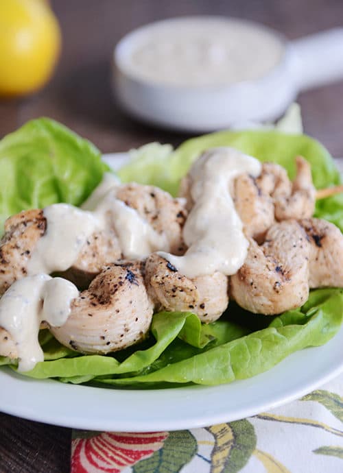 Hummus sauce drizzled grilled chicken skewers laying on lettuce leaves.