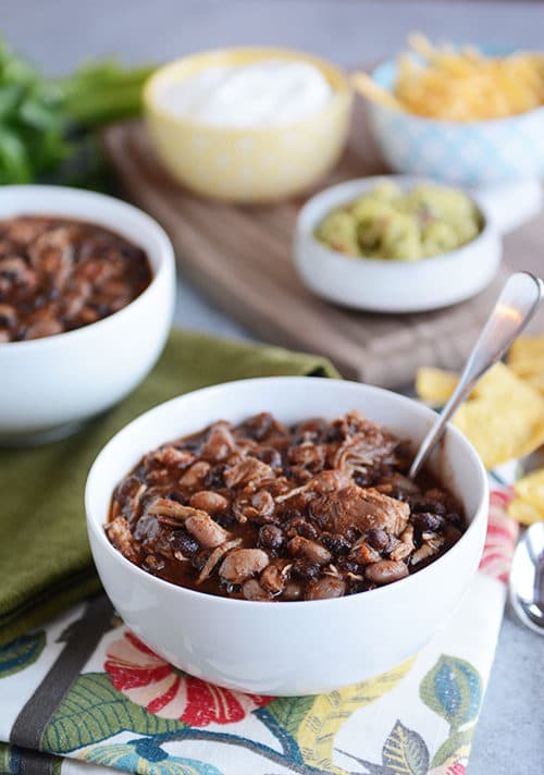 A white bowl of chicken black bean chili with a spoon in it and bowls of toppings in the background.