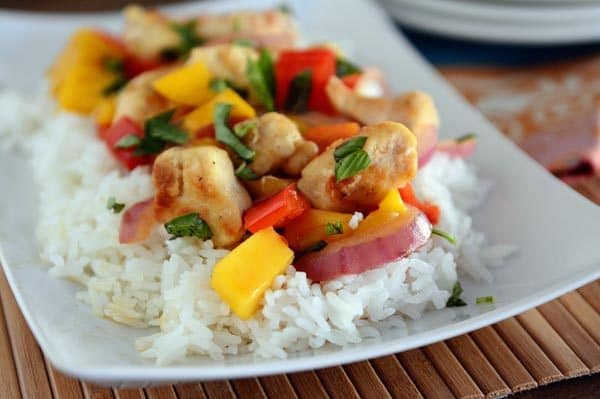 A white platter with cooked white rice topped with a chicken, mango, and onion stir fry. 