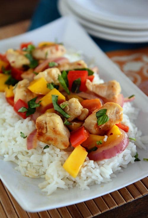 Top view of a white platter full of cooked white rice topped with a chicken, mango, and basil stir fry. 