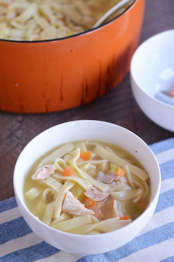 A white bowl of chicken noodle soup with a large pot of the soup behind it.