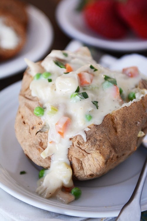 A baked potato on a white plate split open and filled with chicken pot pie sauce.