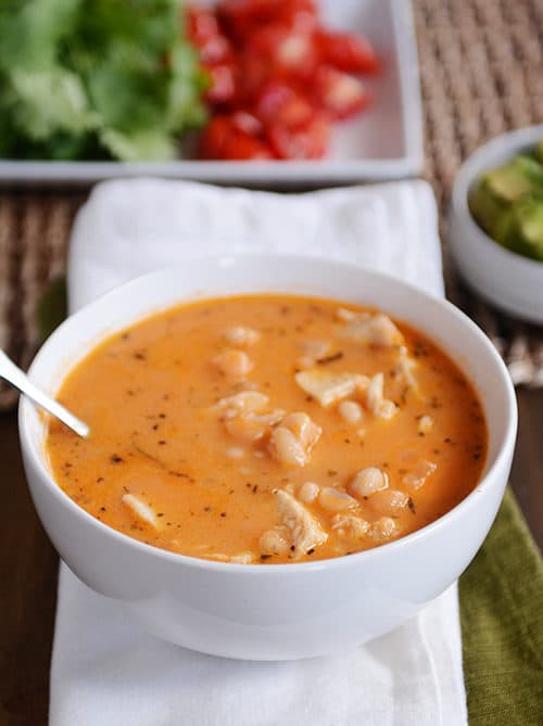 A white bowl full of white bean chicken chili with a tray of lettuce and tomatoes in the background.