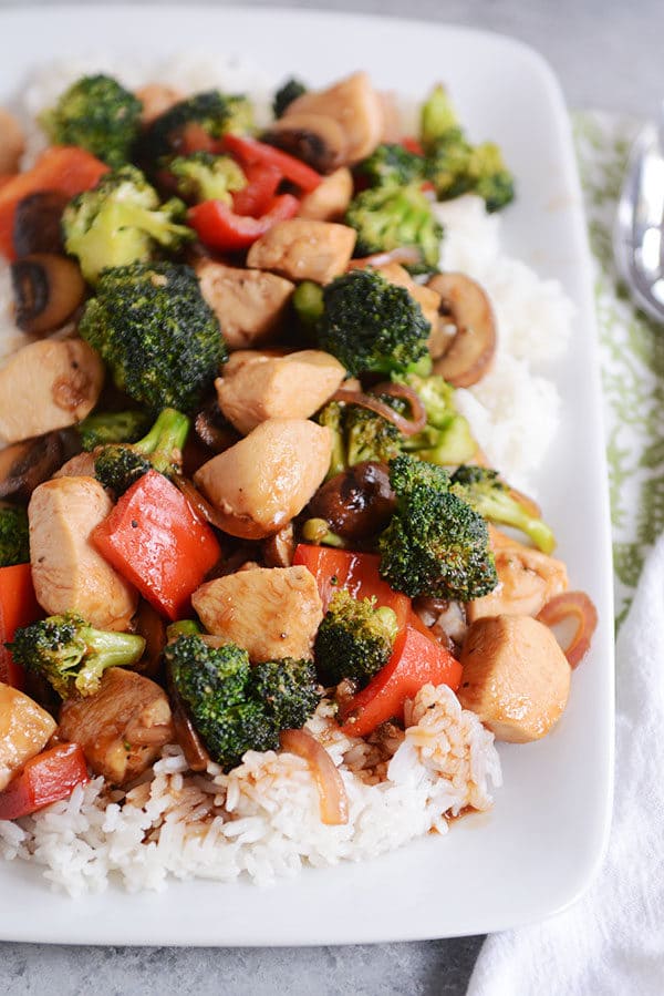 Top view of a white platter of cooked white rice topped with chicken and vegetables.