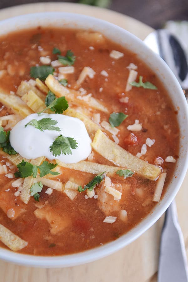 A bowl of red, chicken soup with tortilla strips, sour cream, and cilantro speckling the top. 