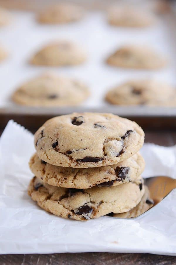 Three chocolate chip cookies stacked on top of each on a metal spatula with a tray of more cookies behind them.
