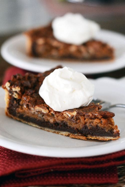 A white plate with a slice of chocolate caramel pecan pie with a dollop of whipped cream.