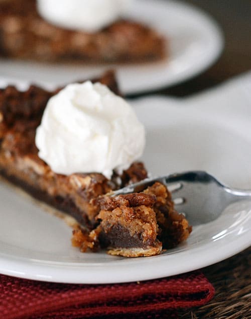 A slice of chocolate caramel pecan pie with a dollop of whipped cream on top and a fork taking a bite out.