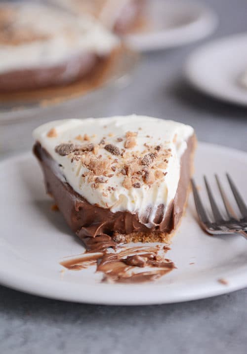 A piece of whipped cream topped chocolate pudding pie with a bite eaten on a white plate.
