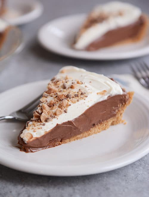 A slice of chocolate pudding pie with graham cracker crust and whipped cream topping on a white plate.