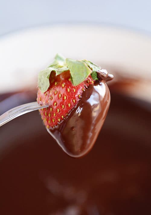 A fork holding a strawberry half dipped in chocolate fondue, over a pot of chocolate fondue.