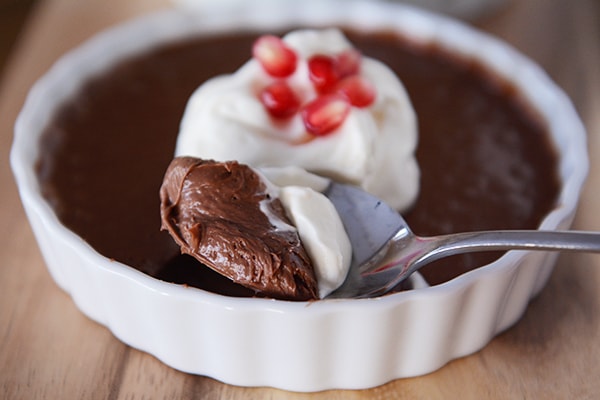 A white ramekin filled with chocolate cream topped with whipped cream and pomegranate seeds, with a spoon taking a bite out.