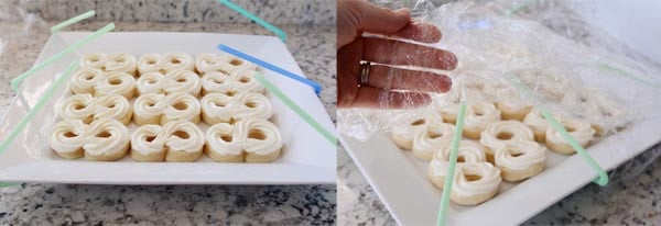 Straws and saran wrap being tented over two white trays of frosted sugar cookies.