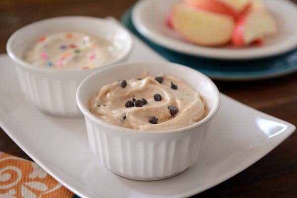 Small white ramekins of funfetti dip and cookie dough dips side-by-side with sliced apples in the back.