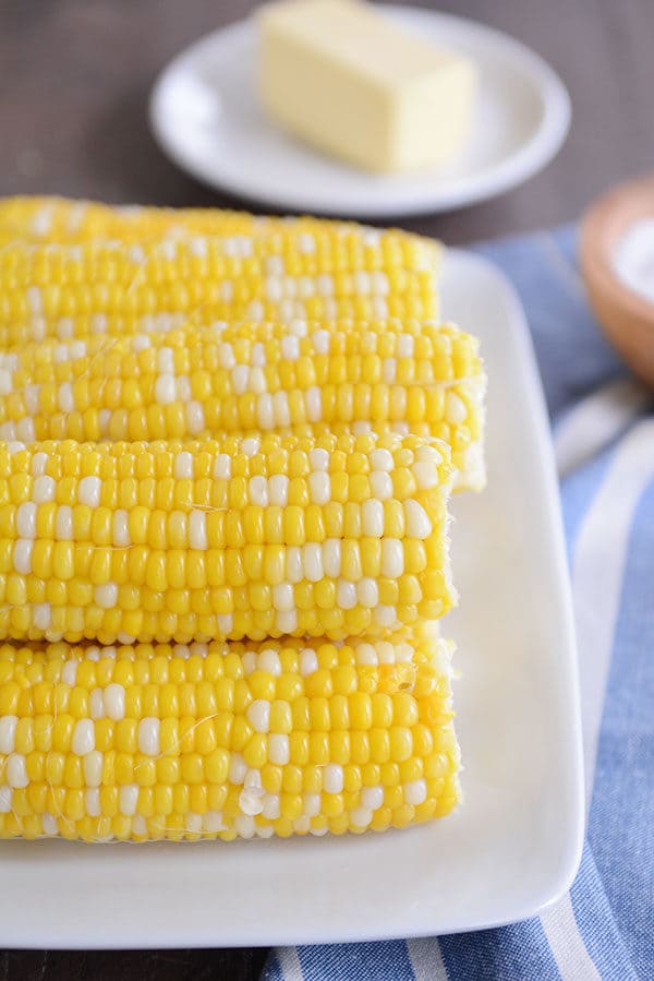A stack of cooked corn on the cobs on a white plate with a plate of butter behind it.
