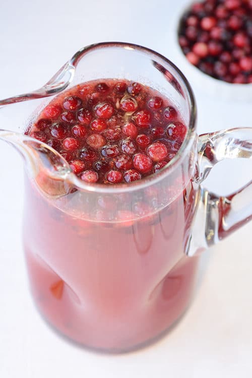 A pitcher full of red punch with cranberries floating on top, and a bowl of cranberries in the background. 