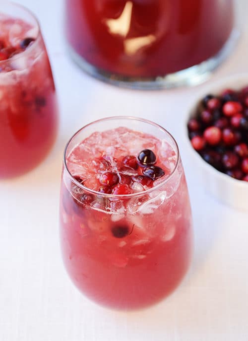 A clear glass full of red punch and floating cranberries on top, with a bowl of cranberries on the side and another glass of punch on the side.