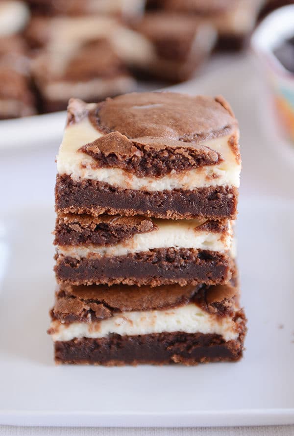 Three cream cheese chocolate brownies stacked on top of each other on a white plate.