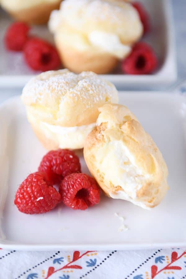 Bite taken out of cream puff on white plate with raspberries.