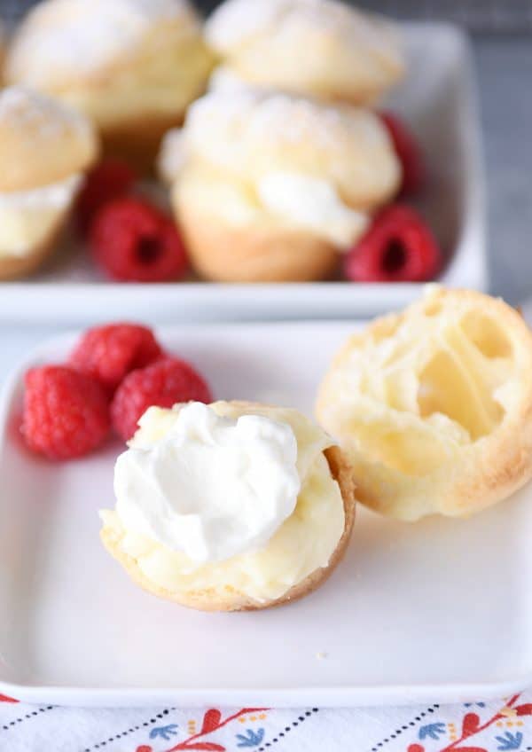 Cream puff split open with cream and raspberries on white plate.
