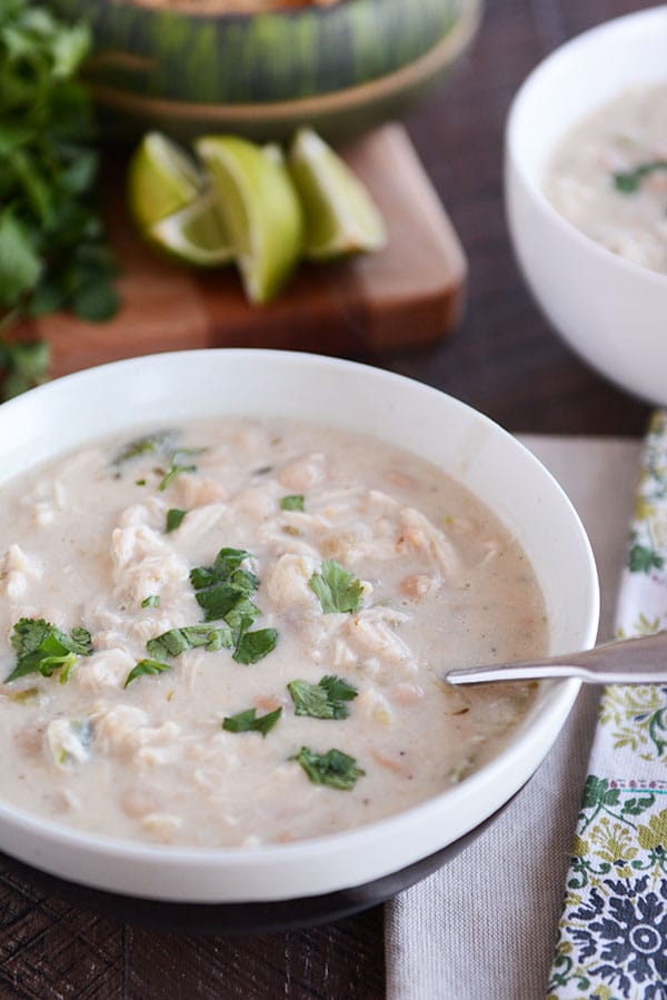 A bowl of white soup with chicken, cilantro, and white beans throughout the soup, and a cutting board with limes and cilantro in the background. 