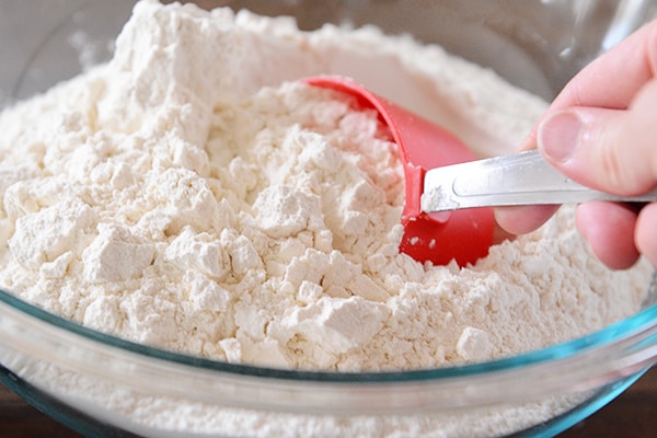 A red measuring cup scooping out flour from a glass bowl.