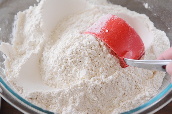 A clear bowl of white flour with a red measuring cup scooping some out.