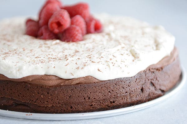 A chocolate torte topped with chocolate mousse, whipped cream, and fresh raspberries on a white plate.