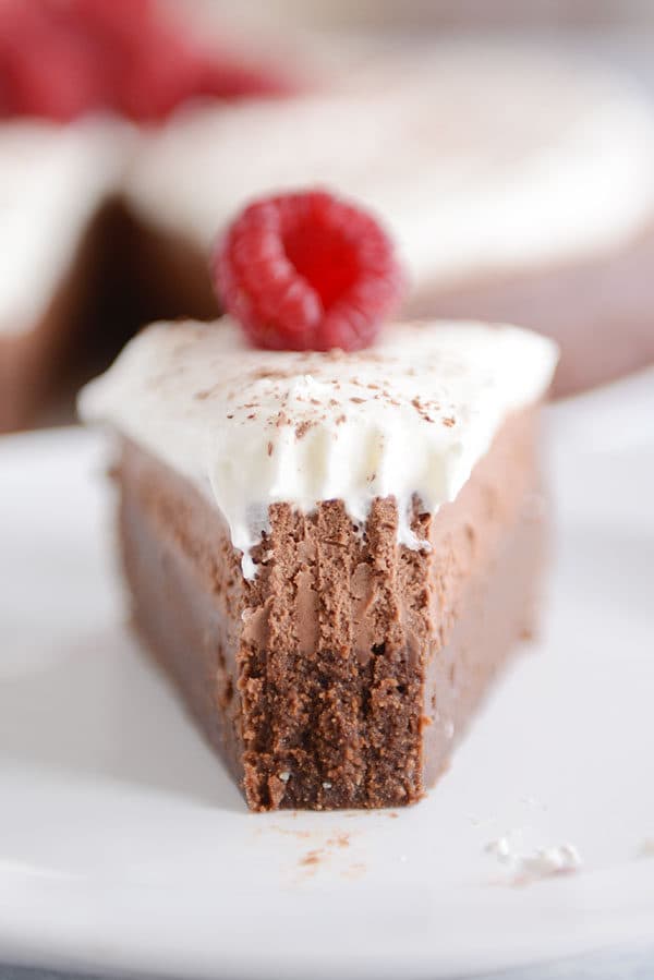 A slice of chocolate torte with a mousse layer in the middle and whipped cream on the top on a white plate.