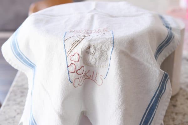 A bowl of bread dough with an embroidered towel covering it.