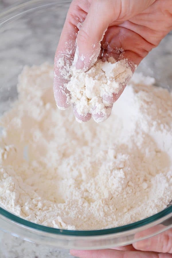 A hand scooping out a mixture of butter and flour from a glass bowl.