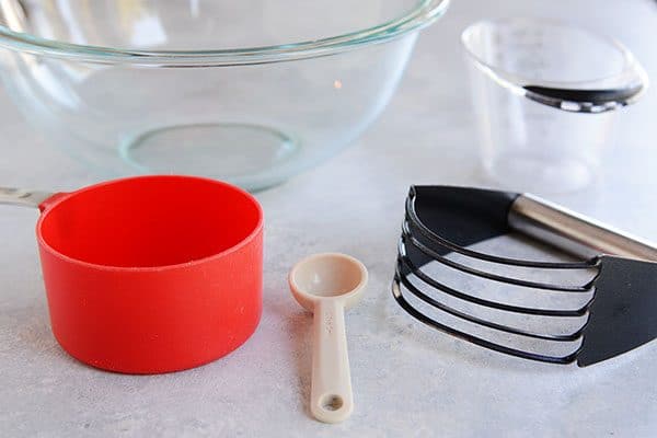A glass bowl, red measuring cup, teaspoon, pastry blender, and liquid measuring cup.