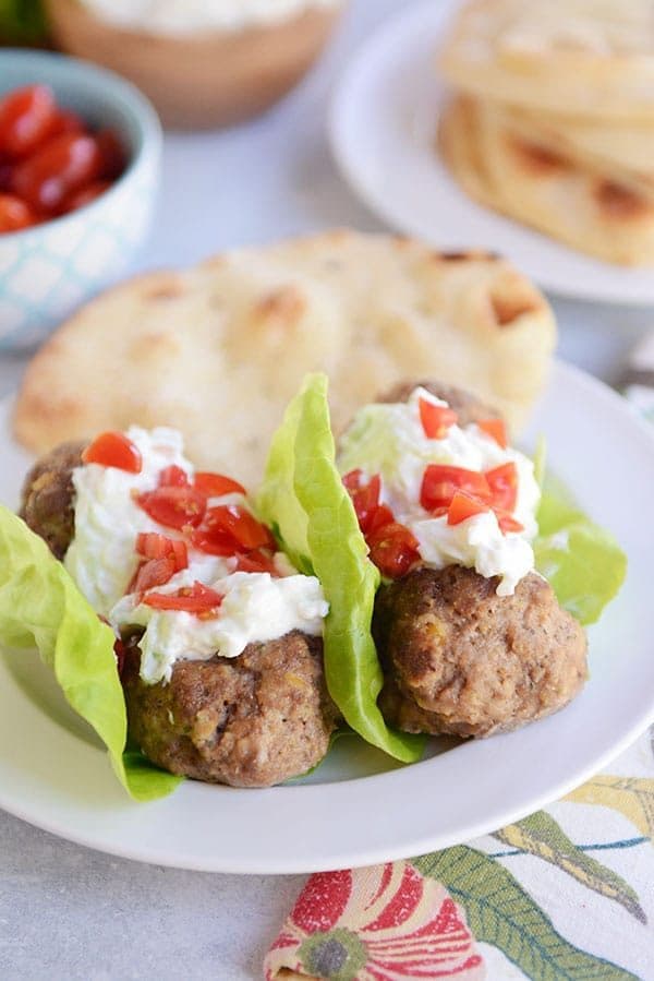 A white plate of falafel meatball lettuce wraps topped with tzatziki sauce and chopped red peppers next to a flatbread.