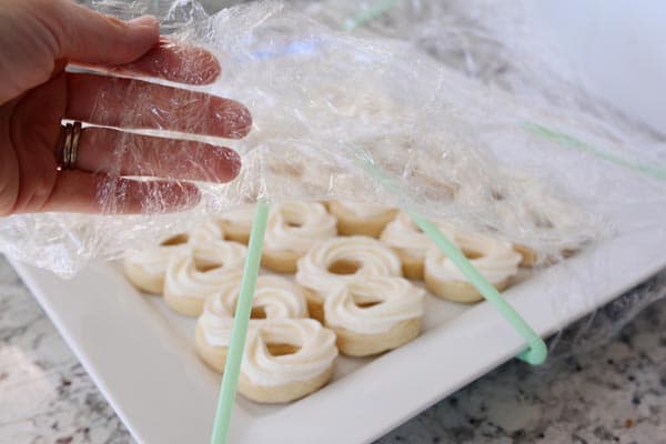 Someone lifting up the side of a saran wrap to show a platter of frosted sugar cookies. 