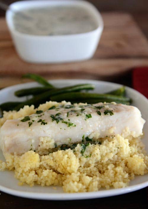 A white plate with cooked couscous topped with a cooked fish fillet and green beans. 