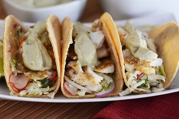 Three corn tortilla broiled tilapia tacos with toppings lined up side-by-side on a white platter.