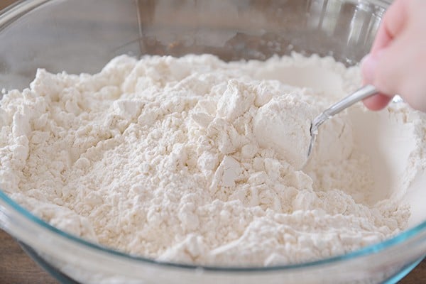 A spoon stirring a clear bowl of white flour. 