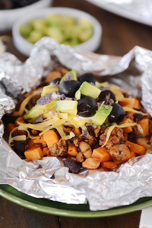 An open tinfoil packet of cooked sweet potato tacos toped with cheese, sour cream, avocado dices, and slices olives.