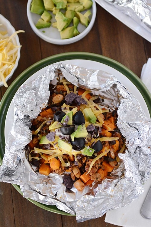 Top view of a tinfoil packet of a cooked sweet potato taco mixture topped with cheese, avocado and sliced olives.