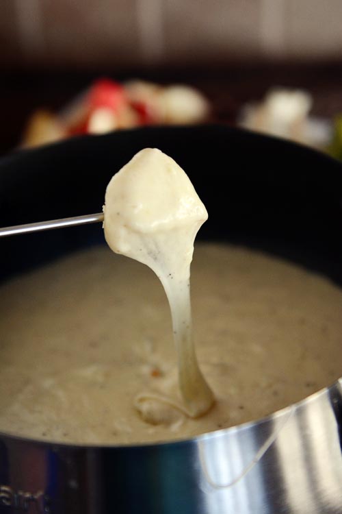 A skewer with a fondue-covered food over the top of a pot of cheesy fondue. 