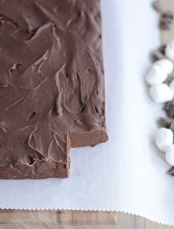 A brick of fudge on white parchment paper, with one square cut out in the corner, and marshmallows and chocolate chips sprinkled on the side of the parchment paper. 