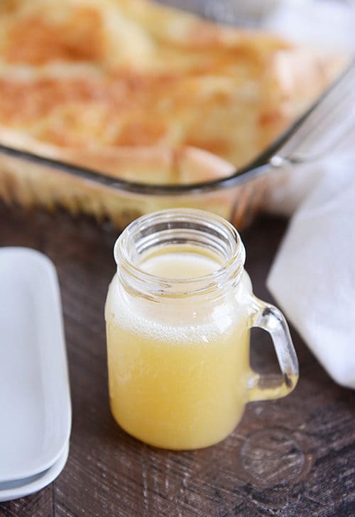 A clear mason jar with a handle full of yellow butter syrup, and a pan of baked german pancakes in the background.