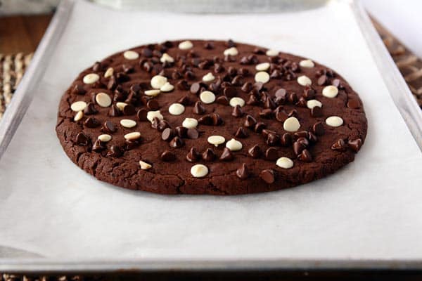 A giant white and chocolate chip topped chocolate cookie on a sheet of parchment paper.