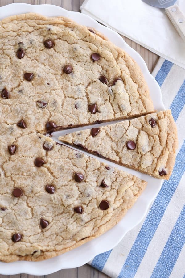 Giant chocolate chip cookie on white platter with slice cut.