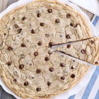 Giant chocolate chip cookie on white platter with slice cut.