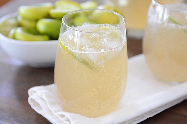 Two glasses of iced limeade in front of a bowl of sliced limes. 
