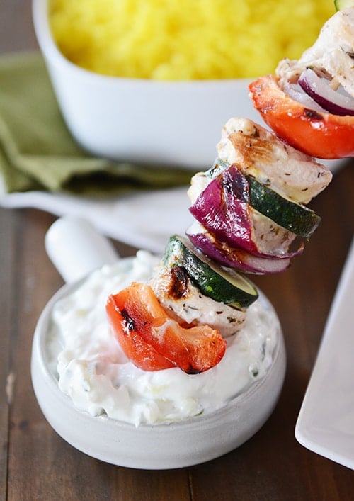 A greek chicken kebab getting dipped into a bowl of sauce. 