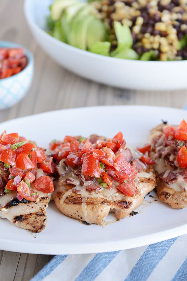 Three grilled chicken breasts with chopped tomatoes on top, and a salad with black beans and corn in the background.