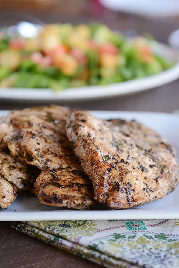 Grilled chicken breasts on a white plate with a green salad behind it.