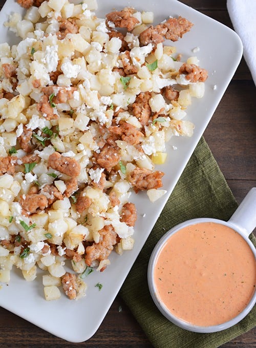 Top down view of a white tray of cubed potatoes and sausage next to a bowl of red pepper sauce.